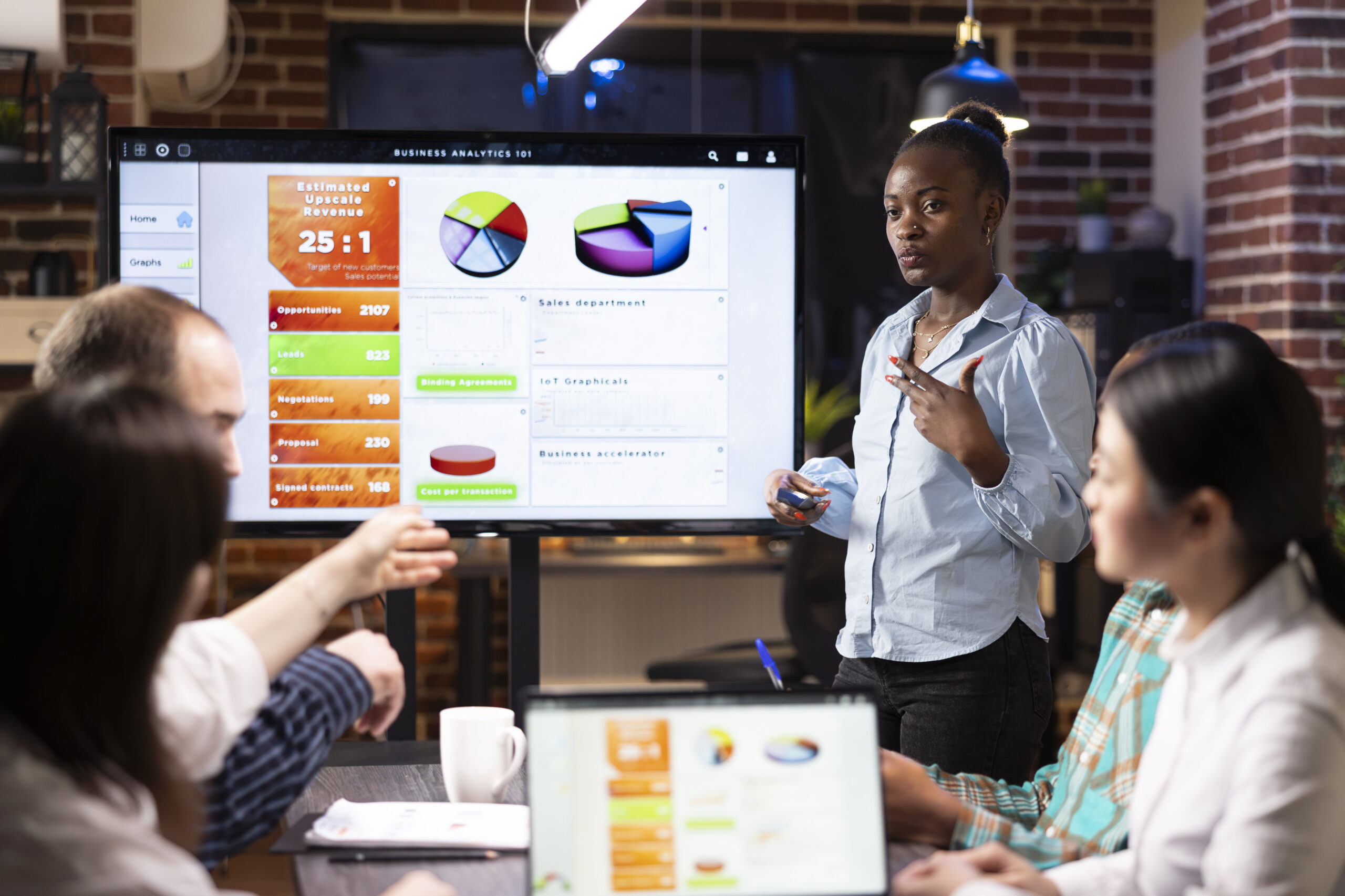 Professional black woman stands near digital monitor, presenting business analytics updates with her colleagues. Multiethnic startup team collaborating in brick wall office, working late at night.